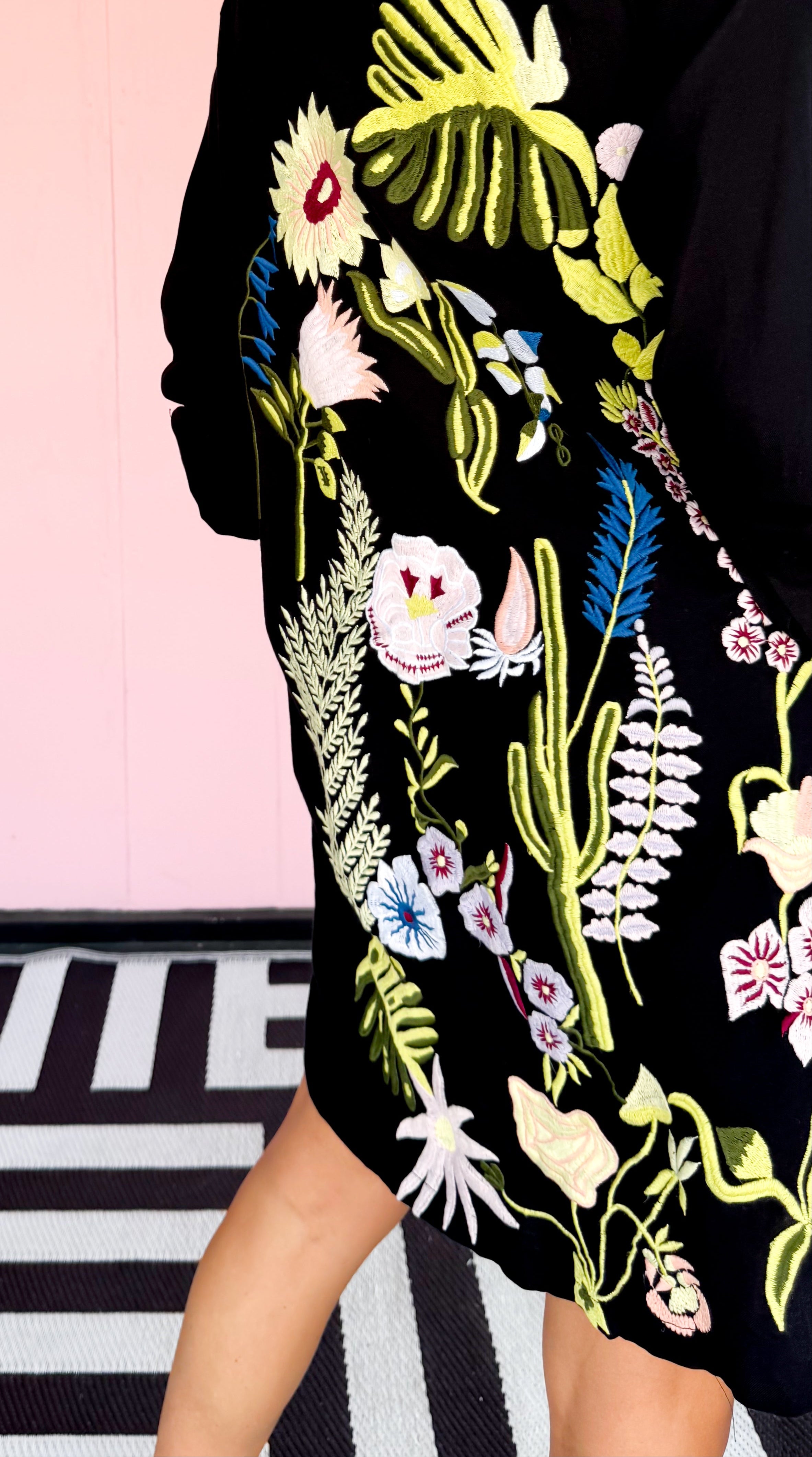 Person wearing a black dress with colorful floral embroidery on a pink and black striped background