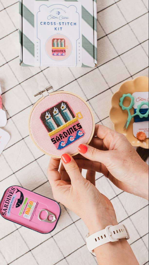 Person holding a cross-stitch kit with 'Sardines' design on a tiled surface.