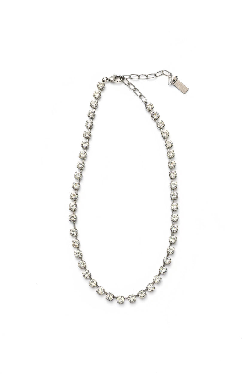 A silver-toned chain necklace with a row of clear round rhinestones, shown with its black retail hangtag on a textured gray fabric background.