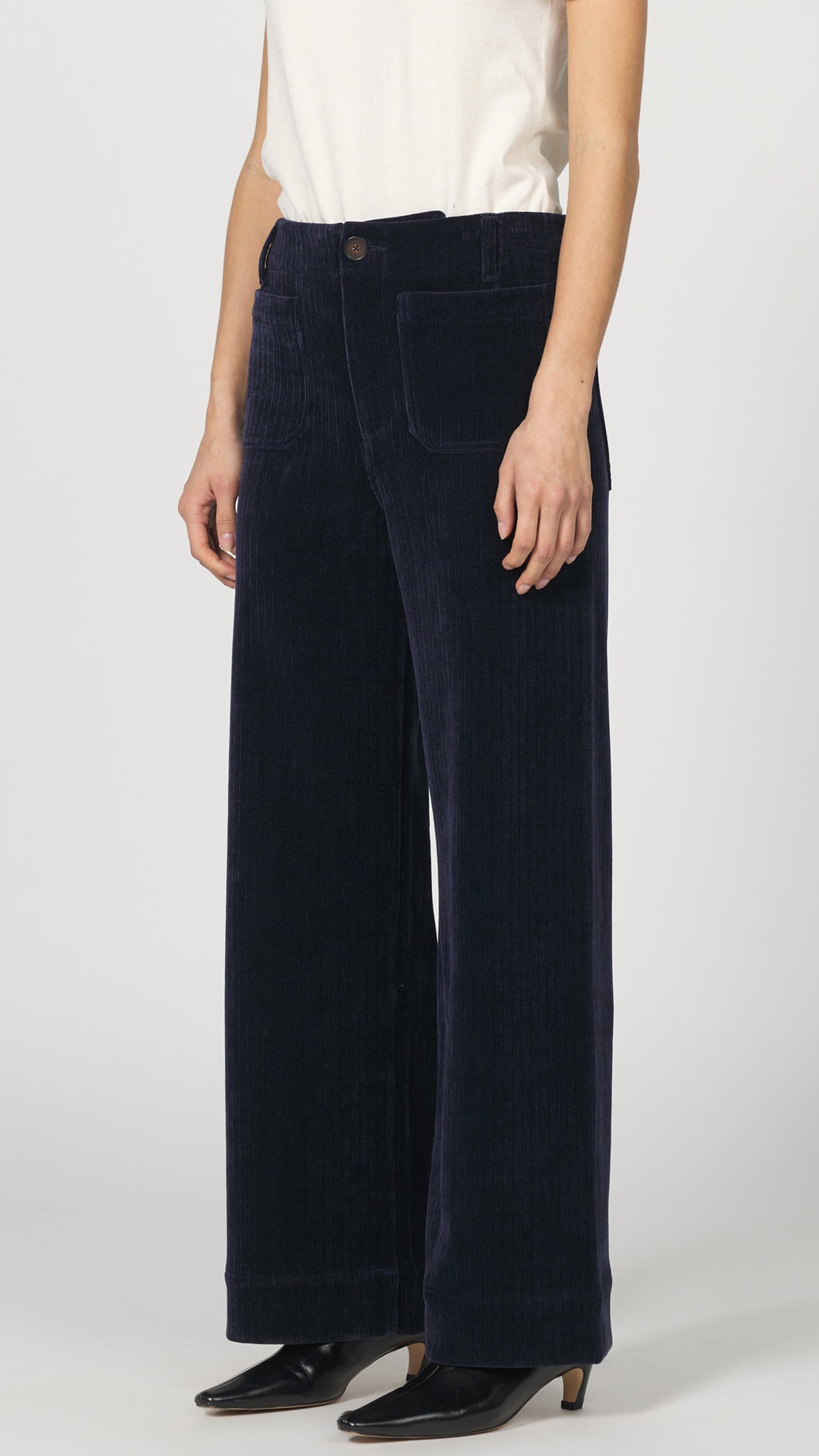 Three-quarter side view of black iris corduroy wide leg pants with a cream tee and black pointed heels