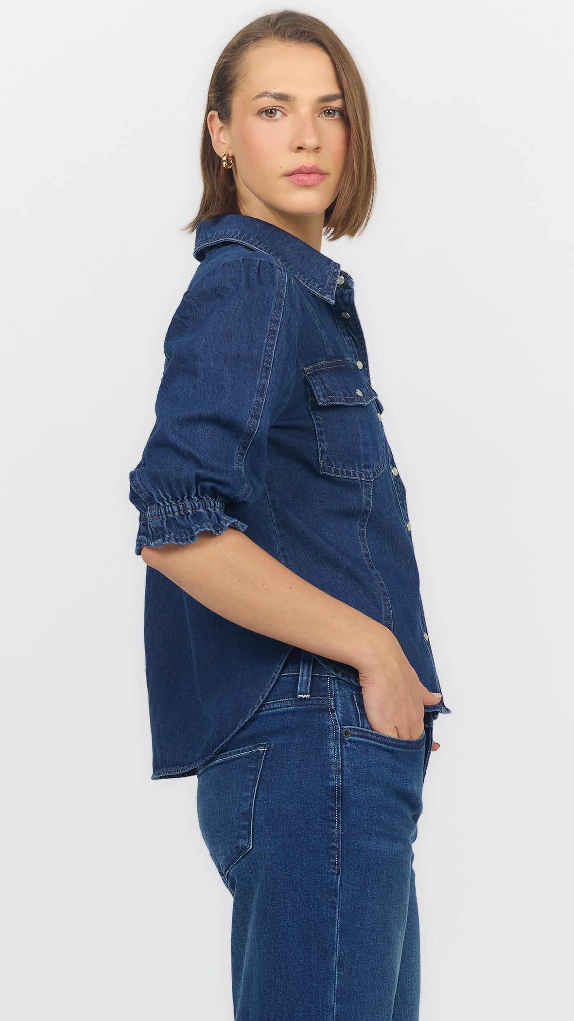 Three-quarter side view of dark cobalt denim blouse showing the ruffled elbow cuff and flap chest pocket, worn with blue jeans