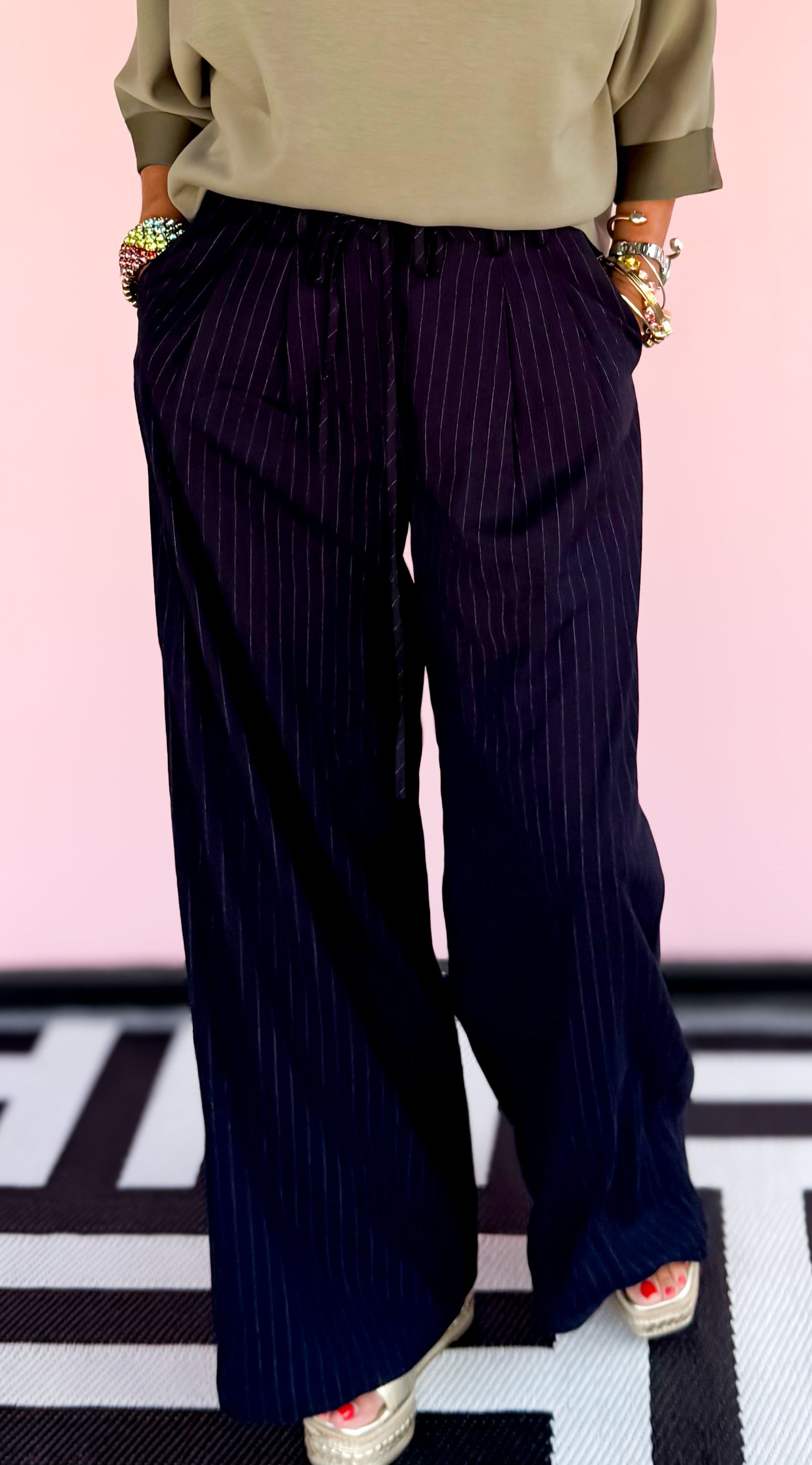 Person wearing an olive green top and dark blue pants against a pink background