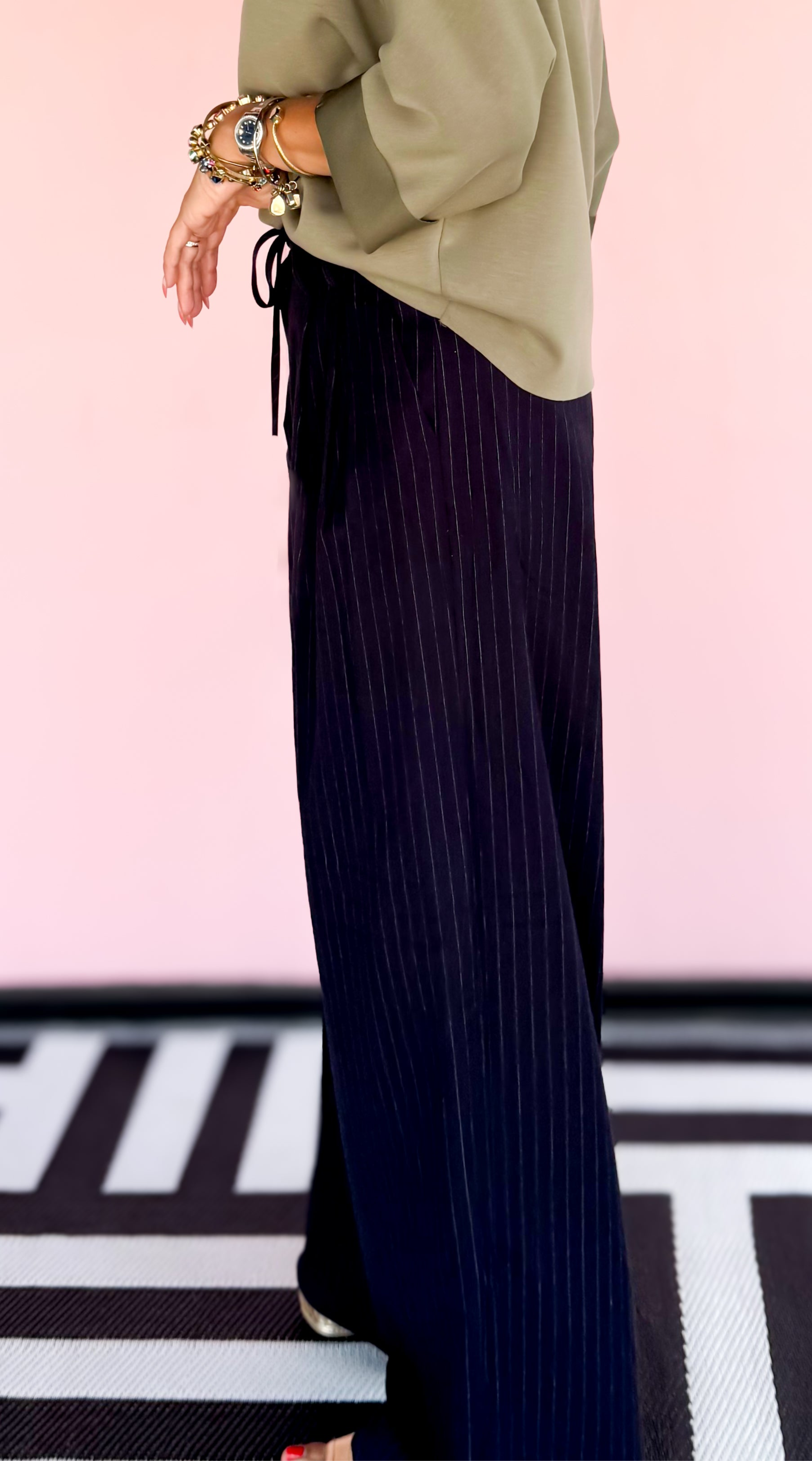Woman wearing a green top and dark pleated pants against a pink background