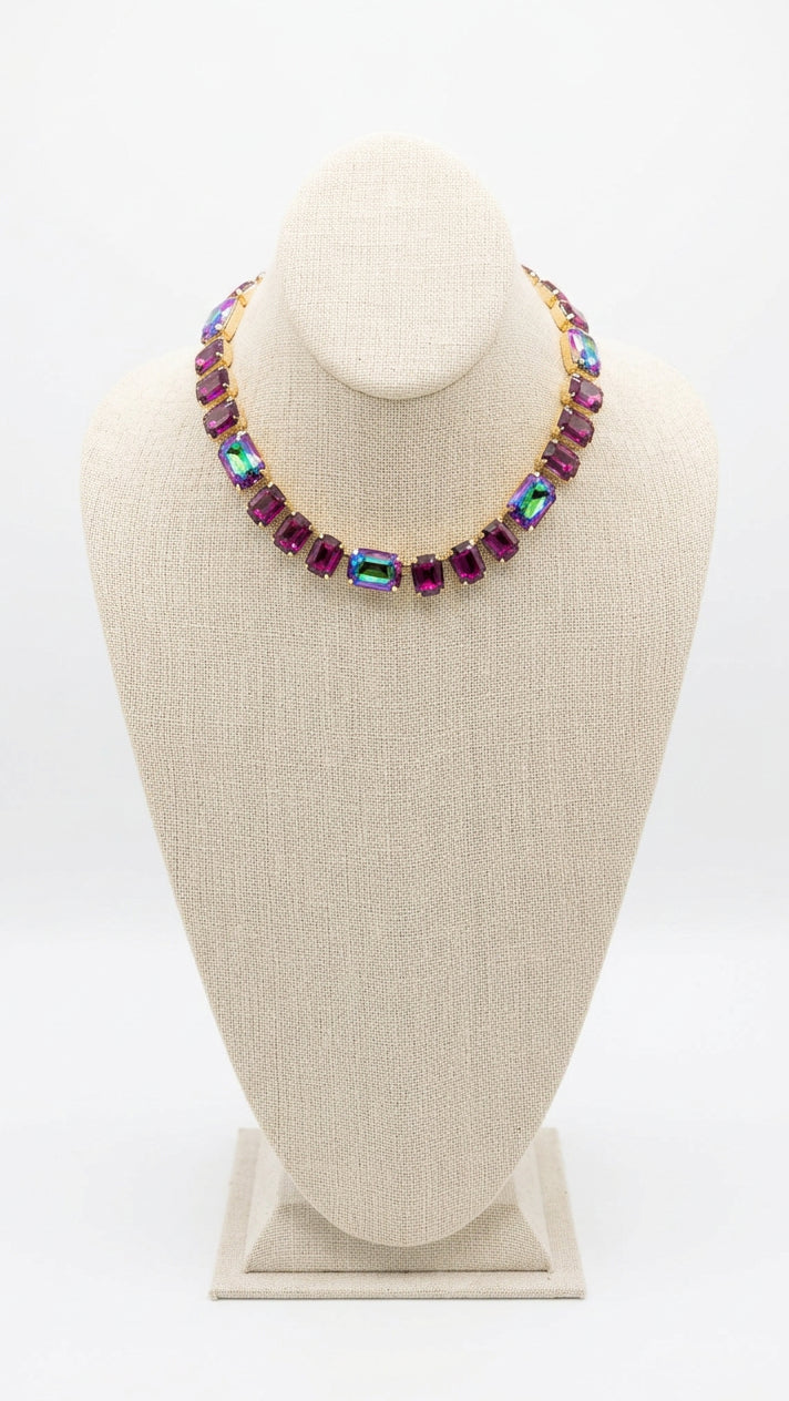 A gold-tone collar necklace with large iridescent magenta and green rhinestones displayed on a beige fabric jewelry bust.
