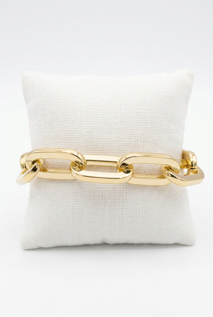 A chunky gold chain link bracelet displayed on a small beige fabric jewelry pillow.
