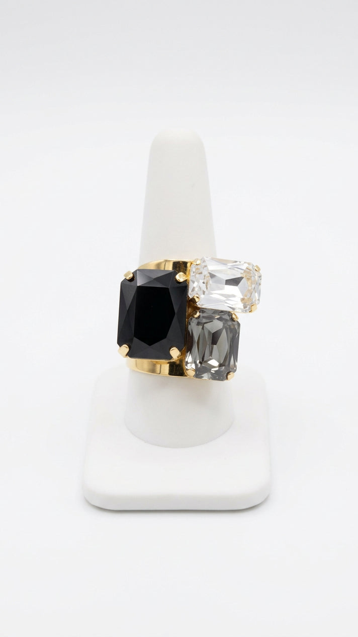 Statement ring with three faceted rectangular rhinestones in black, clear, and smoky gray, arranged asymmetrically in a gold-tone setting on a white ring display.