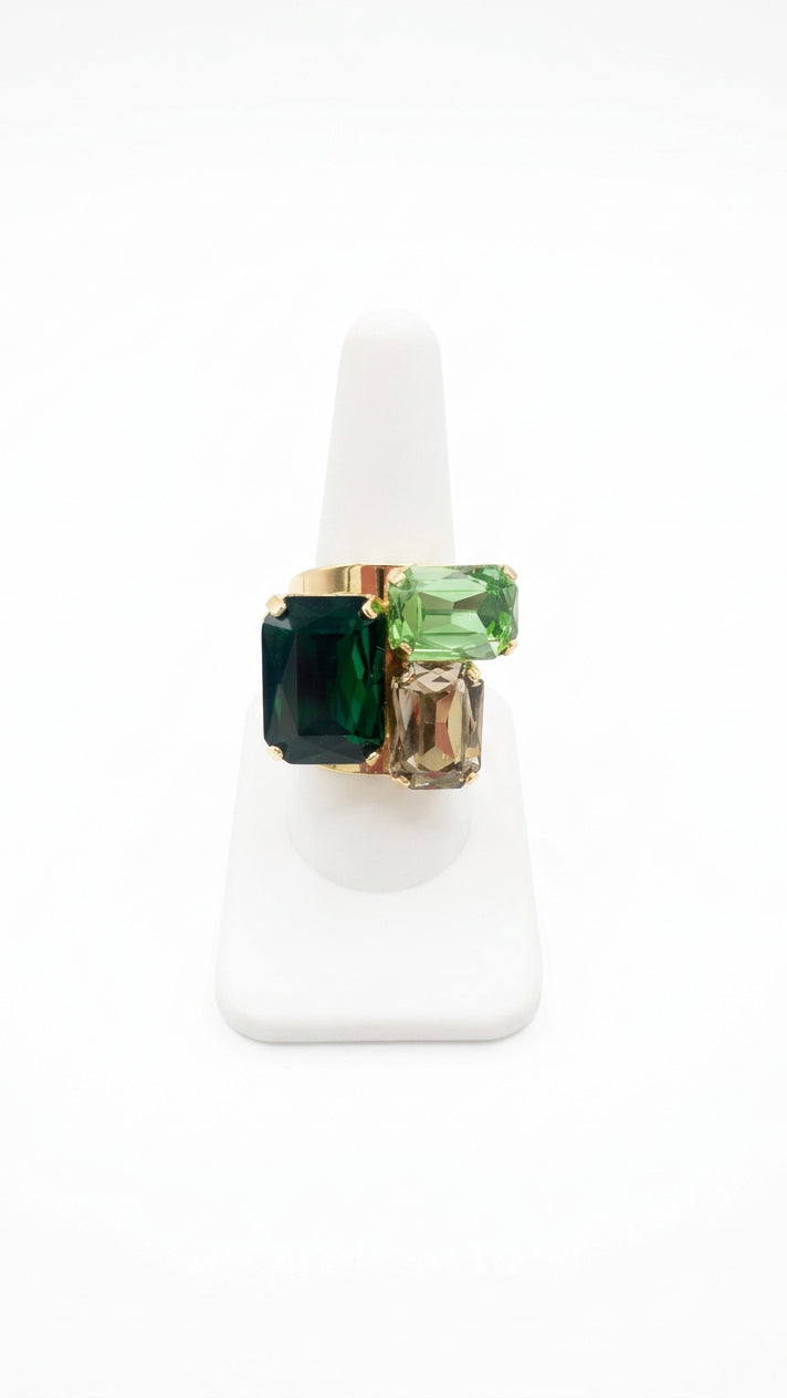 Green statement ring with three faceted rectangular rhinestones in deep green, bright green, and smoky-clear tones, arranged asymmetrically in a gold-tone setting on a white display.