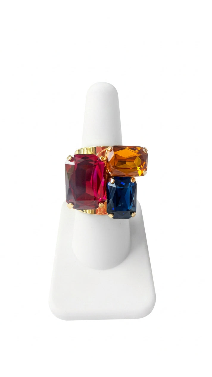 Multicolor statement ring with three faceted rectangular rhinestones in deep pink, amber, and navy blue, arranged asymmetrically in a gold-tone setting on a white display.⠋