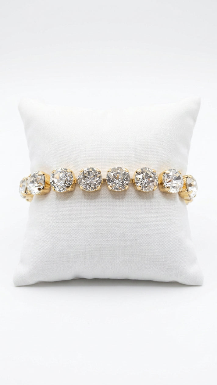 Bracelet with a single row of large, round clear rhinestones in gold-tone settings, displayed around a white jewelry cushion.