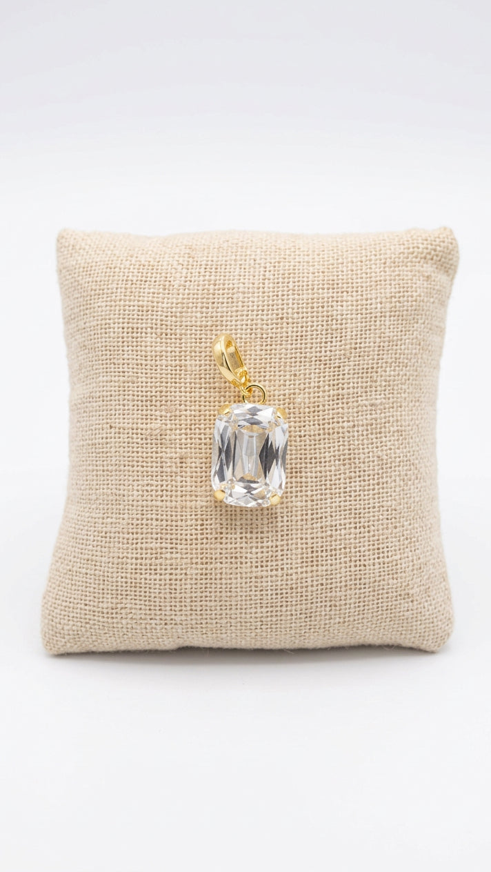 A rectangular clear gemstone pendant with a gold bail, displayed on a small beige fabric cushion over a dark wood surface.
