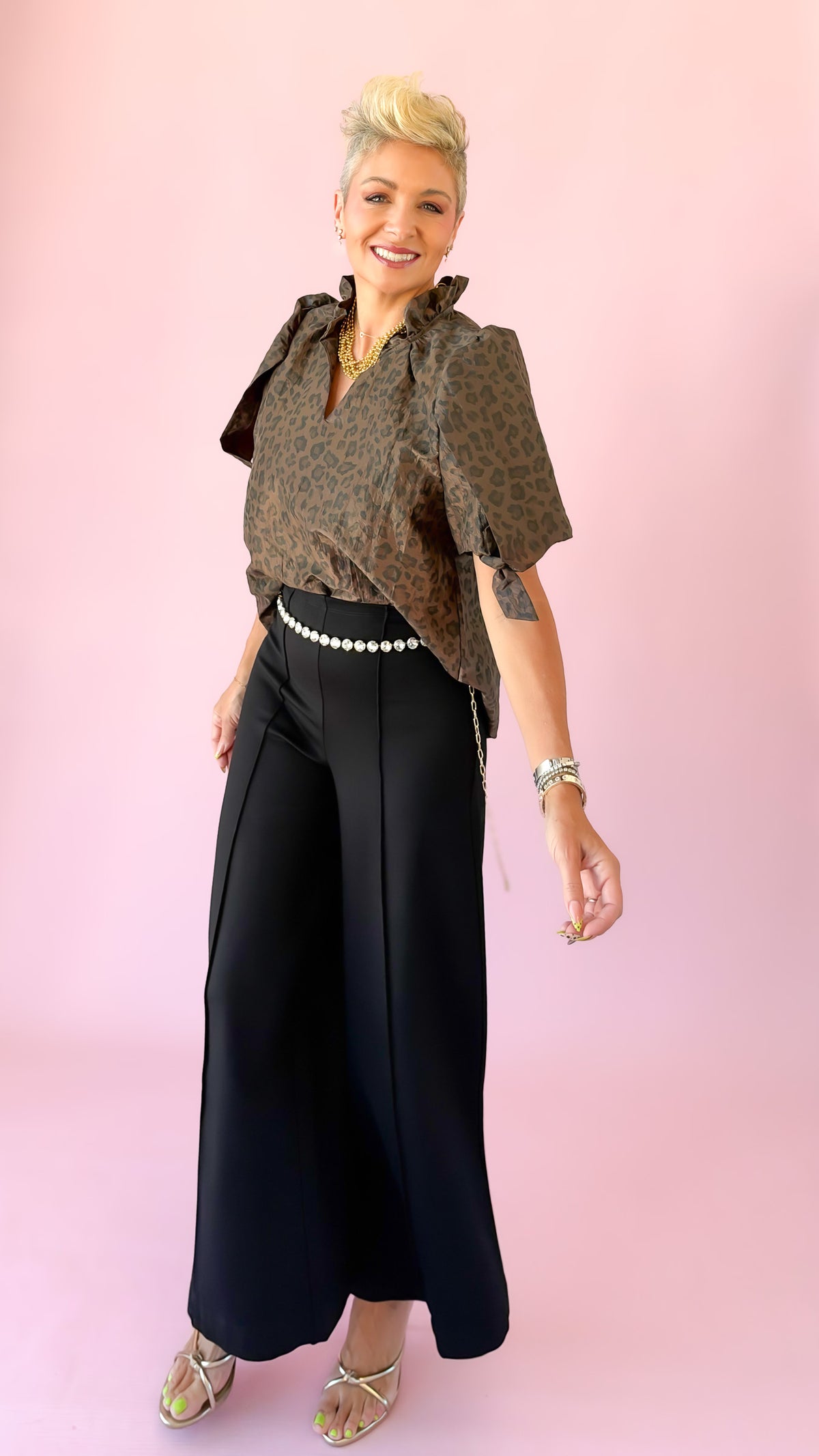 A woman modeling a short-sleeve brown and black patterned blouse with a high ruffled collar, paired with black wide-leg pants and gold heels, standing against a solid pink background.