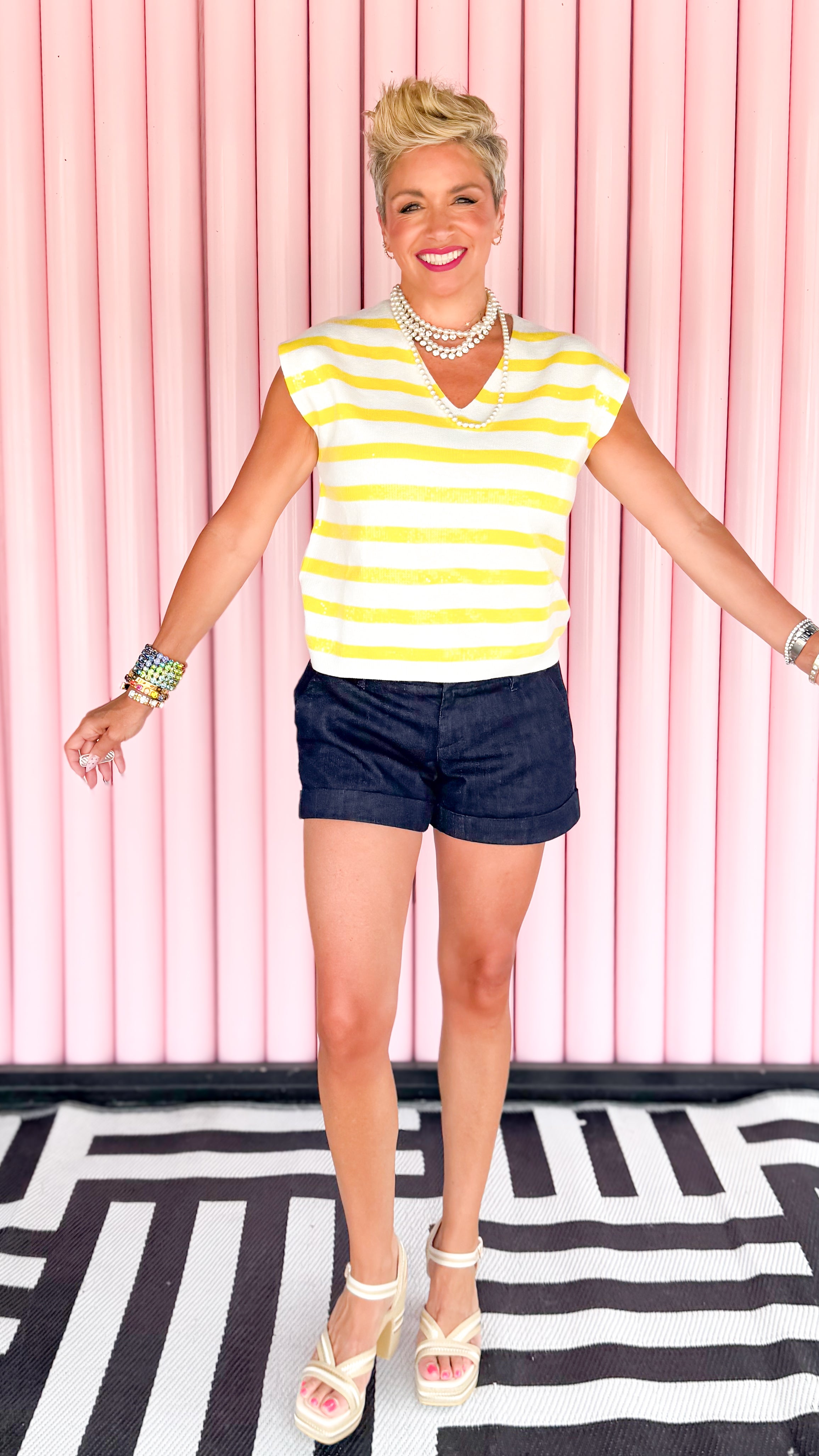 Woman wearing a yellow and white striped shirt with denim shorts against a pink and black geometric patterned background.