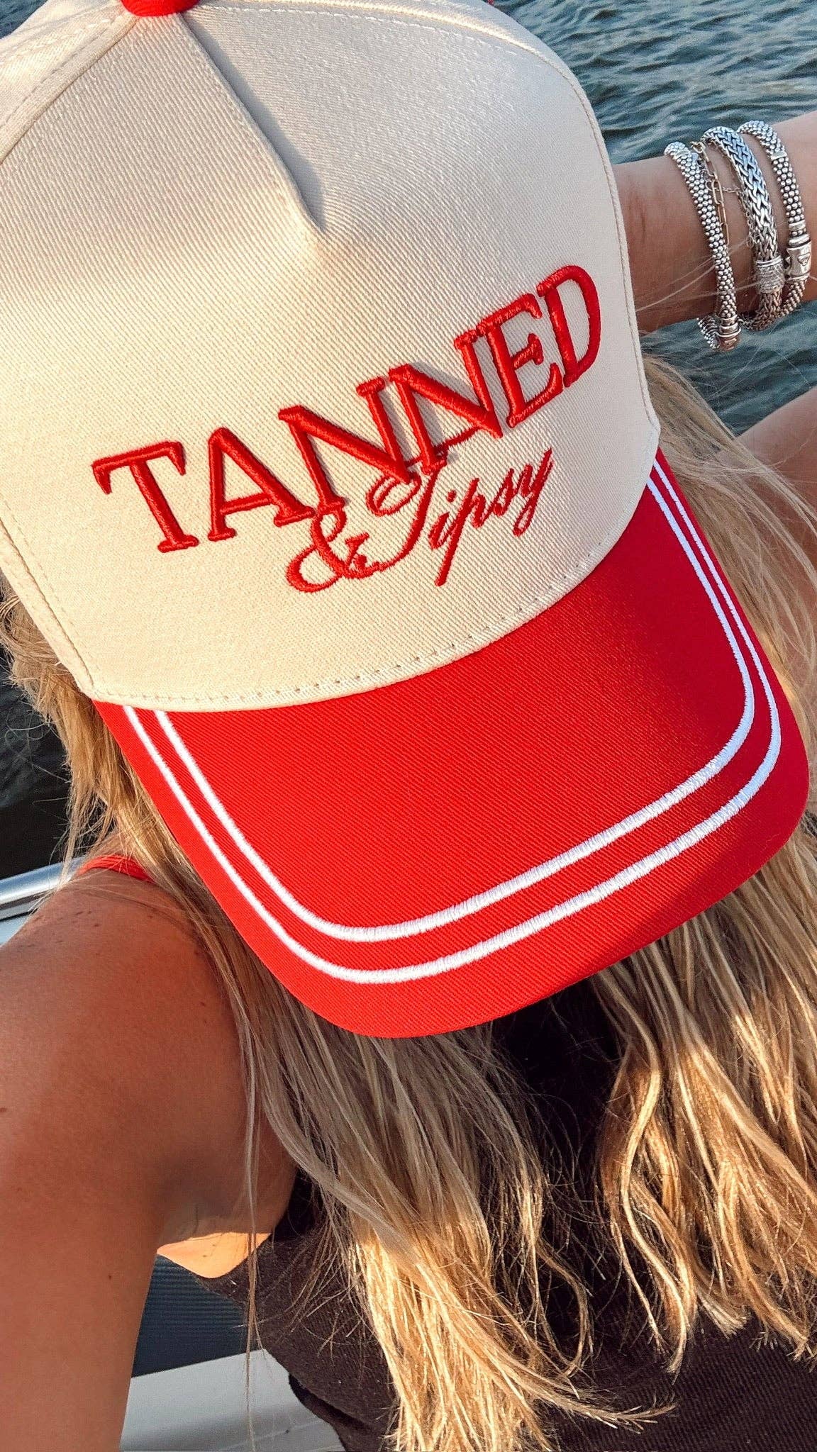 Person wearing a cap with 'Tanned & Tipsy' text on a boat.
