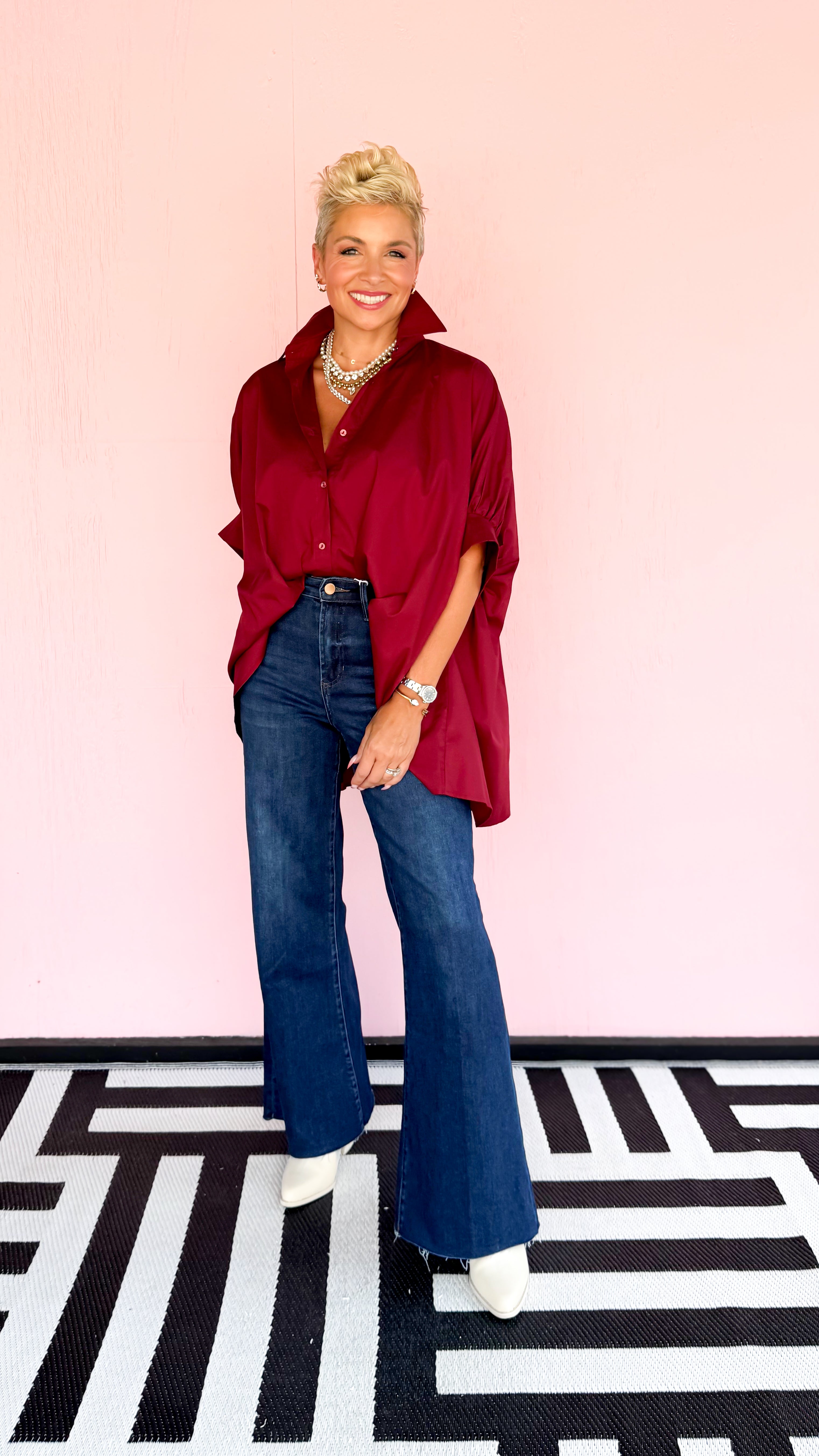 Woman wearing a red blouse and blue jeans standing on a black and white patterned floor with a pink wall.