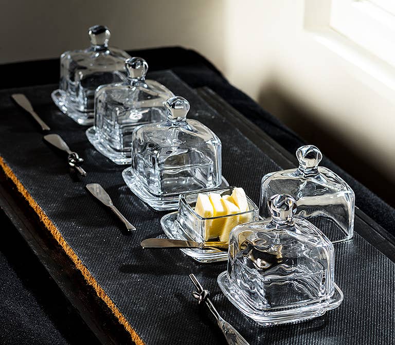 Set of clear glass butter dishes with lids on a dark surface.
