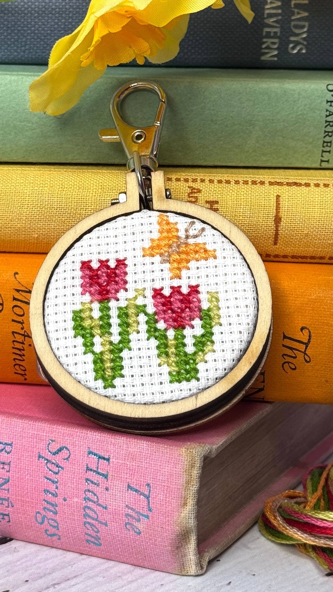 Cross-stitched keychain on a stack of books with yarn and flowers.