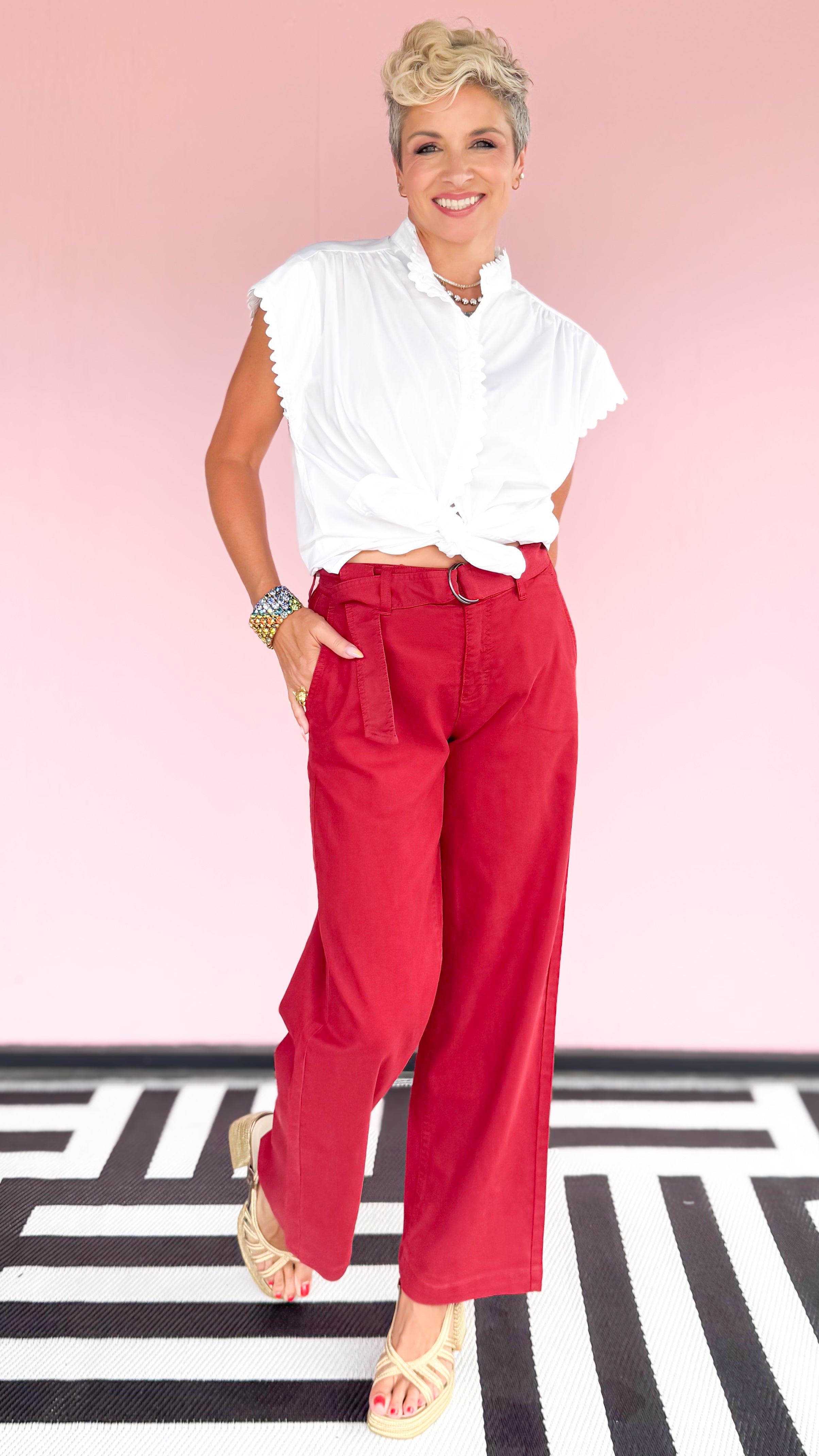 Woman wearing a white blouse and red pants standing on a black and white striped floor with a pink background.