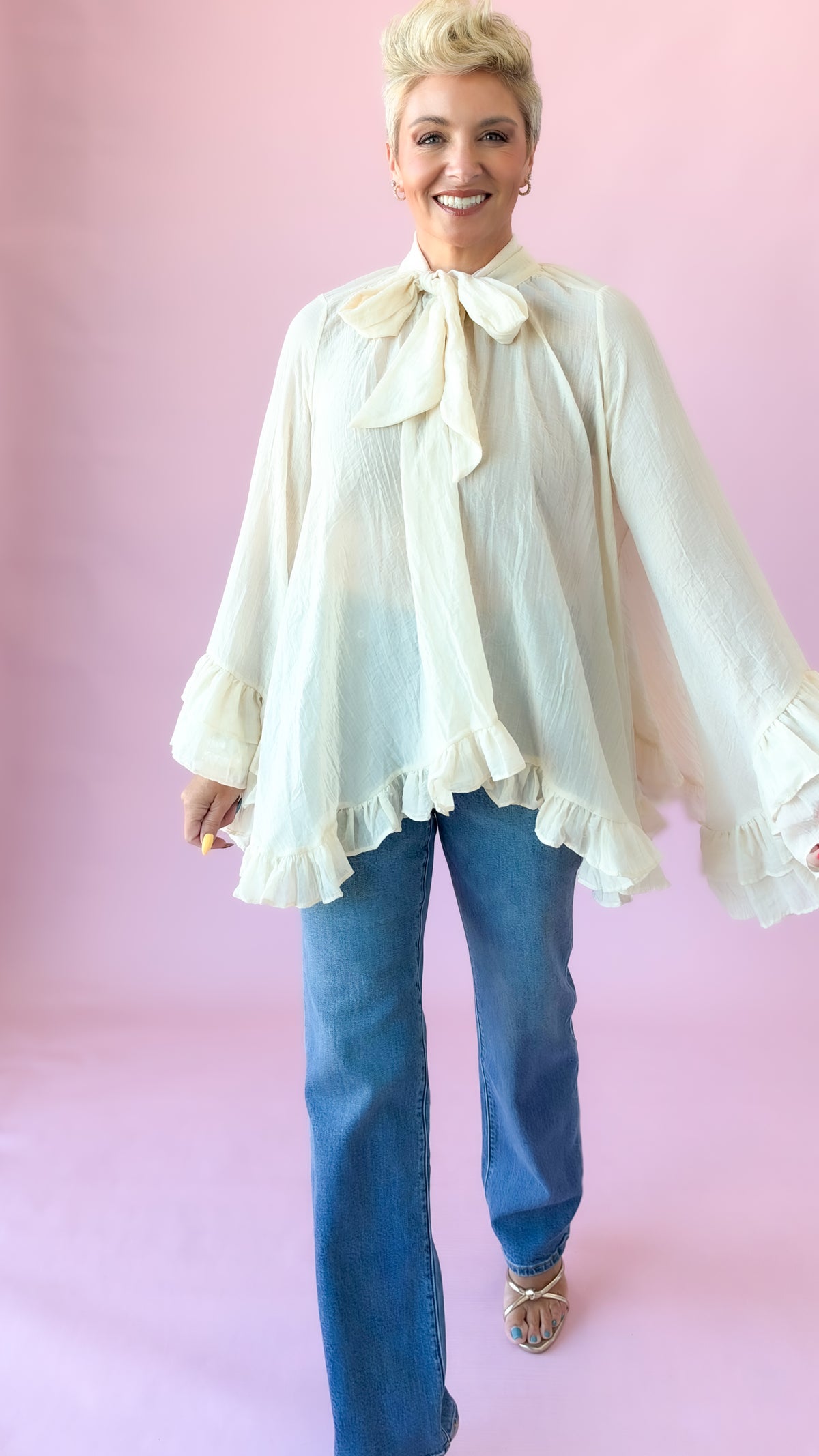 Cream oversized blouse with neck bow and ruffle hem, arms out to show wide sleeves