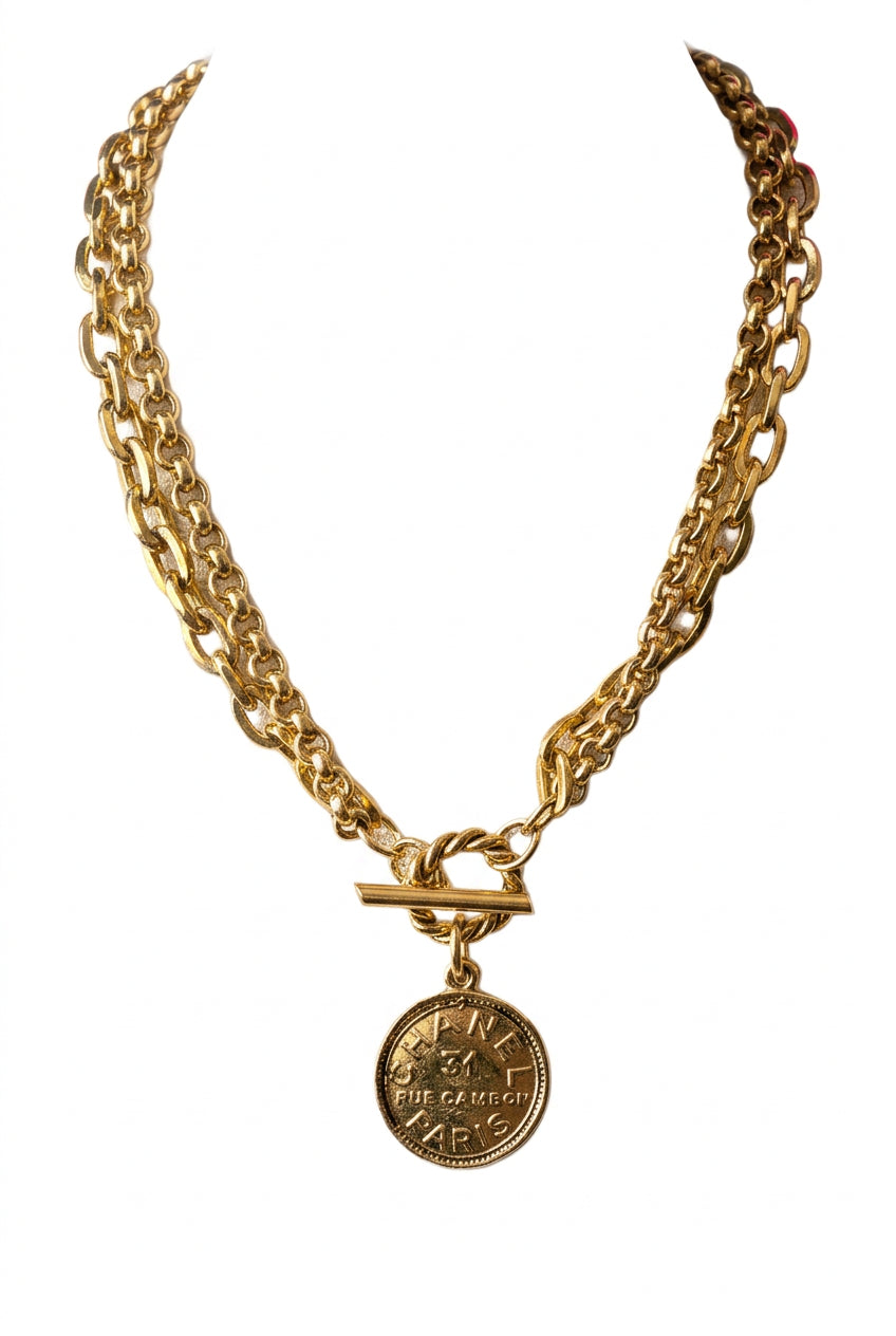 A gold-toned chunky chain necklace with a toggle clasp and round medallion pendant, displayed on a beige fabric jewelry bust.