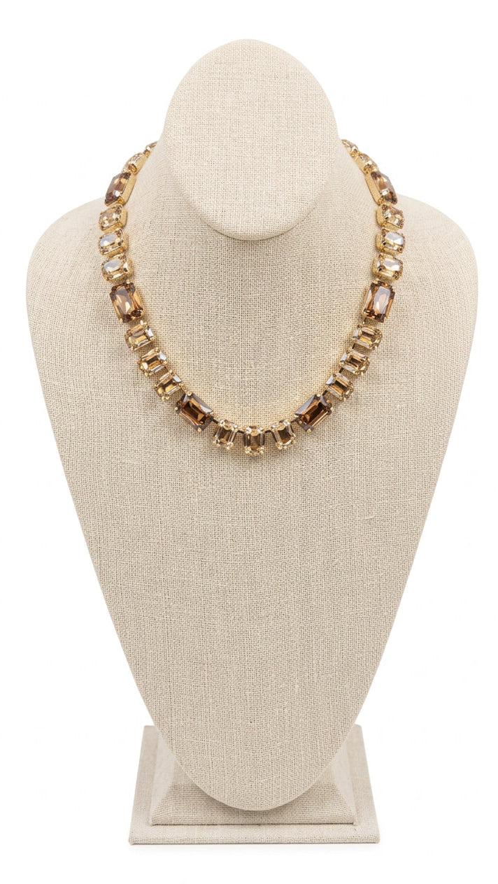 A gold-toned statement necklace with rectangular faceted amber-brown crystals, displayed on a beige fabric-covered jewelry bust. 