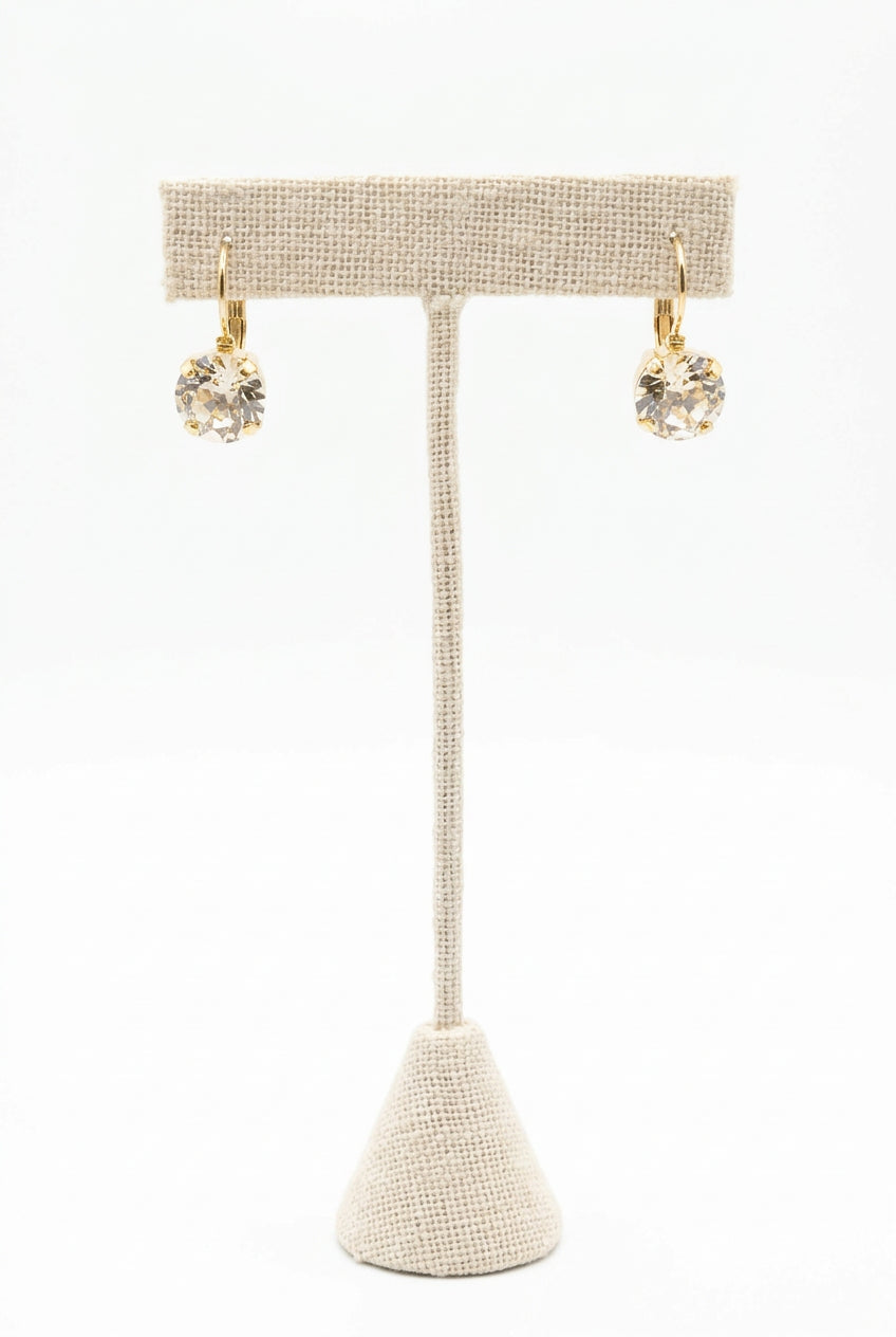 Gold-tone drop earrings with round, faceted clear stones in prong settings, hanging from a beige fabric-covered jewelry stand against a white background.