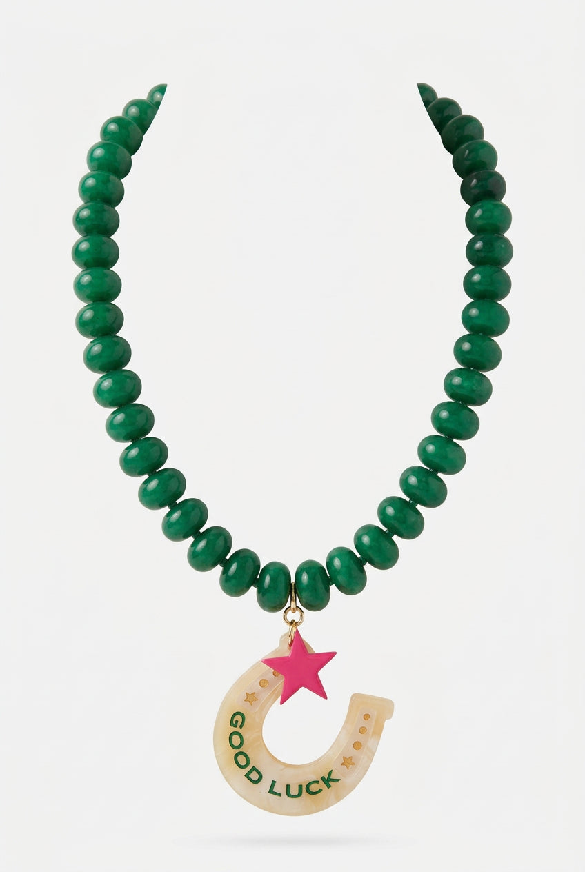 A green beaded necklace with a crescent moon pendant containing the words 'GOOD LUCK' and a pink star, displayed on a beige fabric mannequin bust.