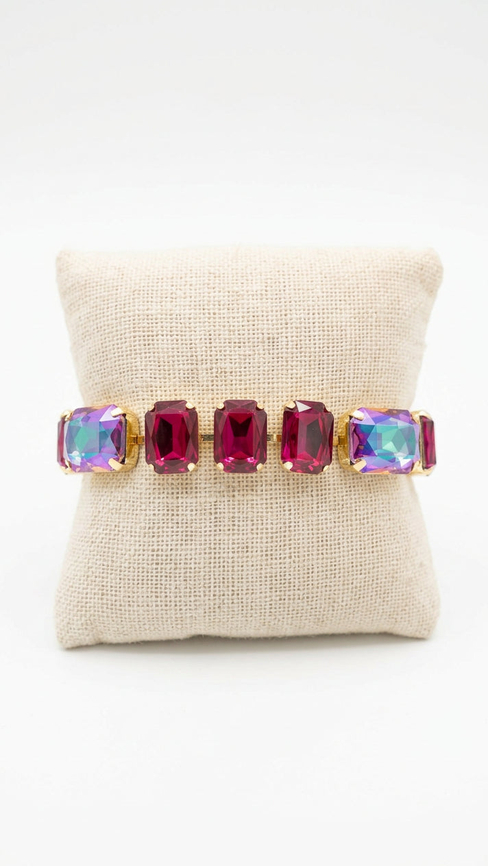 A gold-tone bracelet with five rectangular magenta-purple crystals, displayed on a beige fabric cushion.