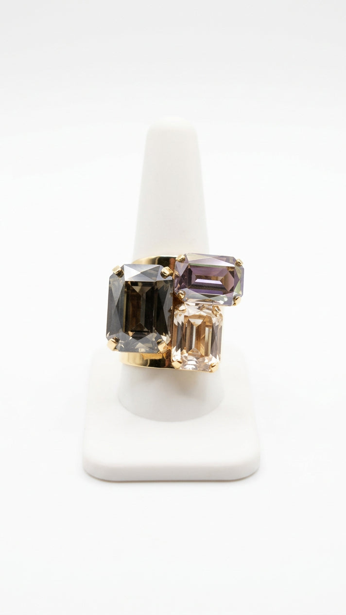 Gray statement ring with three faceted rectangular rhinestones in smoky gray, pale lavender, and champagne tones, arranged asymmetrically in a gold-tone setting on a white display.⠋