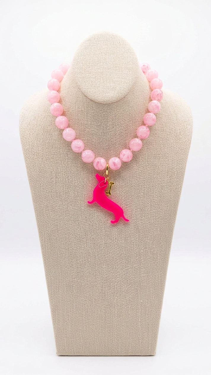 Pale pink beaded necklace with a hot pink dachshund pendant and small gold-tone bone charm, displayed on a beige fabric necklace bust against a white background.⠋