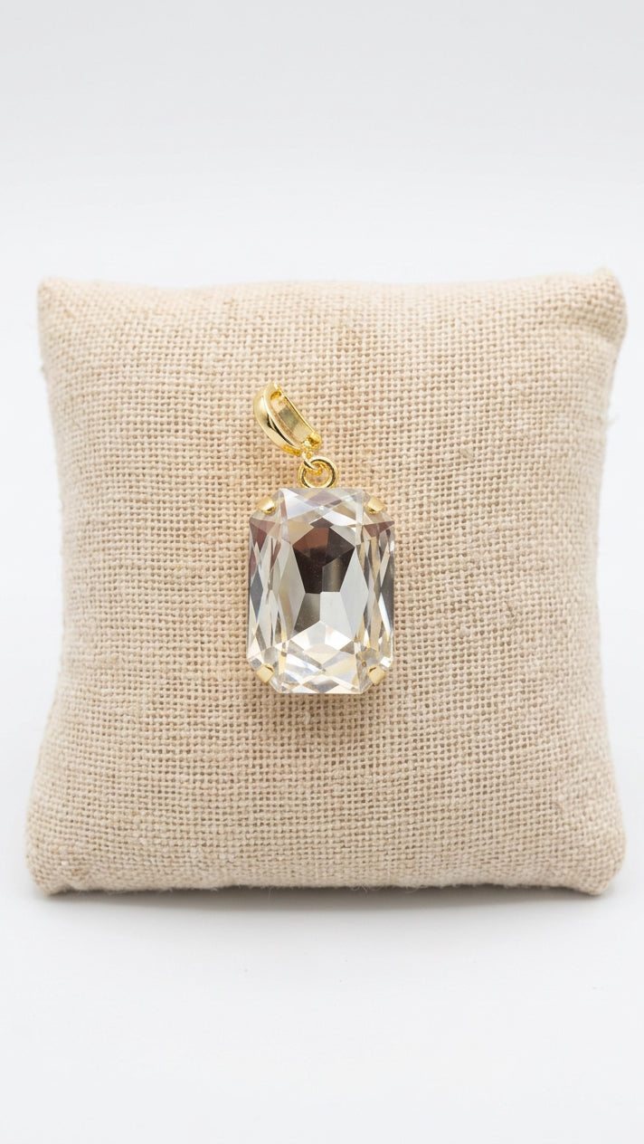 A rectangular clear faceted gemstone pendant with a gold bail, displayed on a small beige fabric cushion over a dark wooden surface.