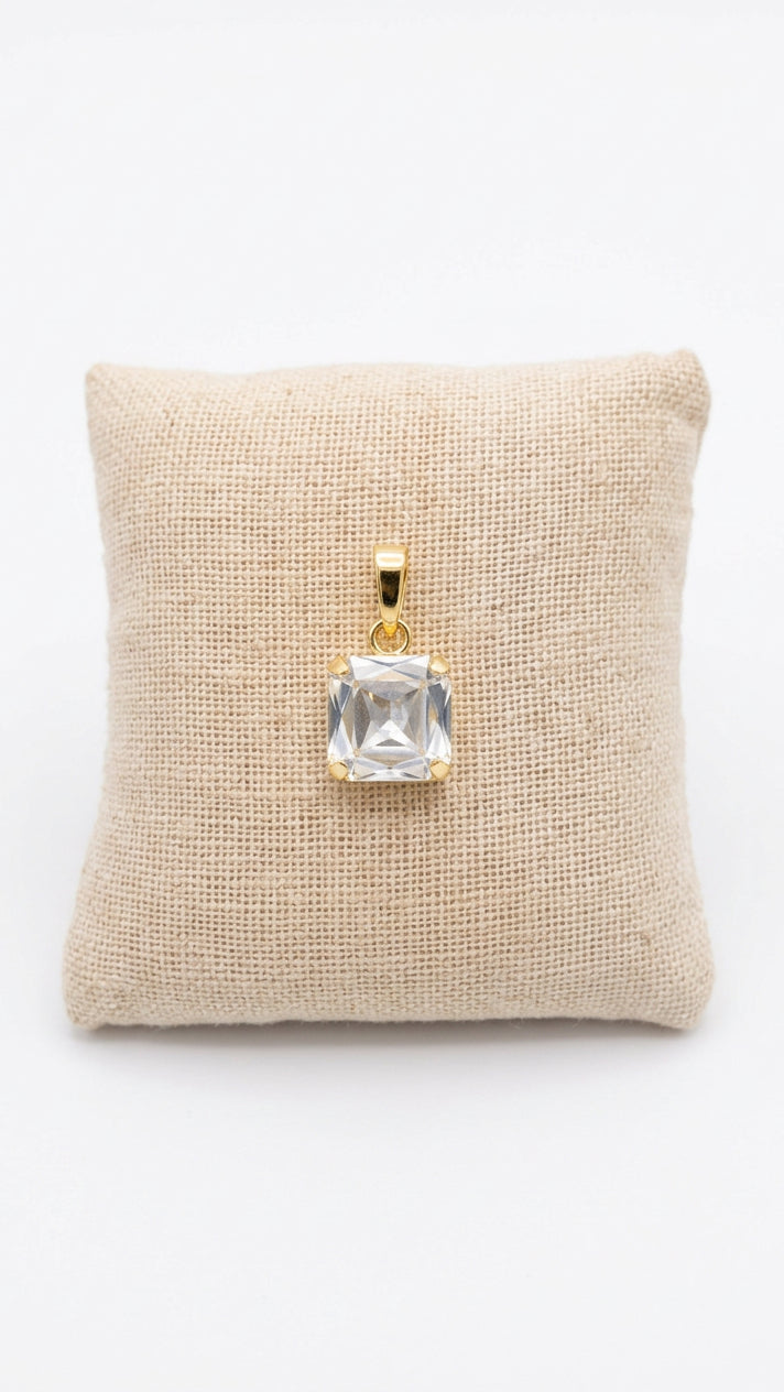 A square clear gemstone pendant with a gold setting, displayed on a small beige fabric cushion .