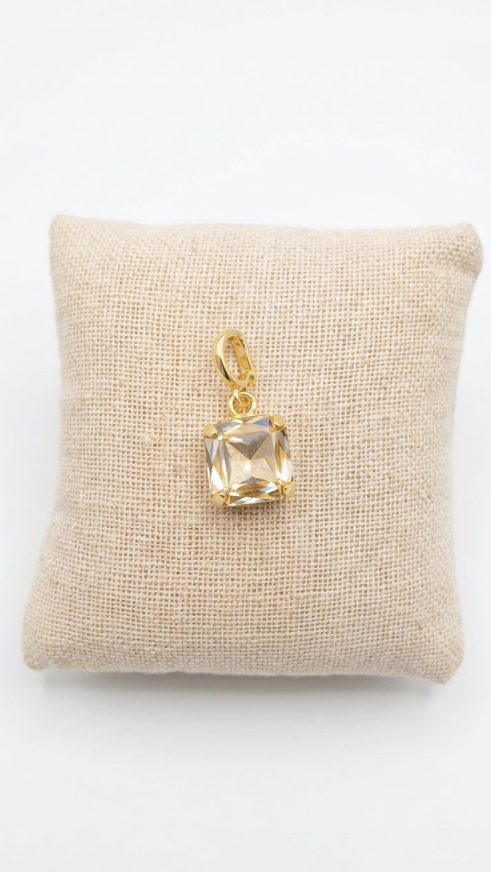 A gold pendant with a square-cut clear gemstone displayed on a small beige fabric cushion.