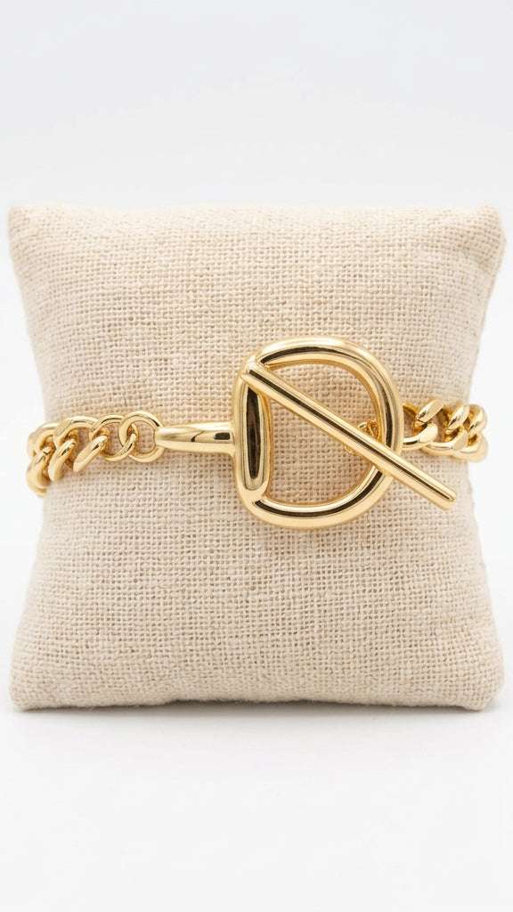 A gold-tone chain bracelet with a large interlocking D-shaped toggle clasp, displayed on a beige fabric pillow held by a hand. 