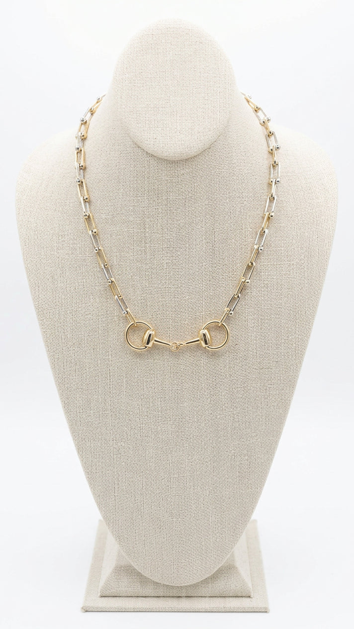 A gold-toned metal link necklace with two interlocking circular ring charms, displayed on a beige fabric jewelry bust. 