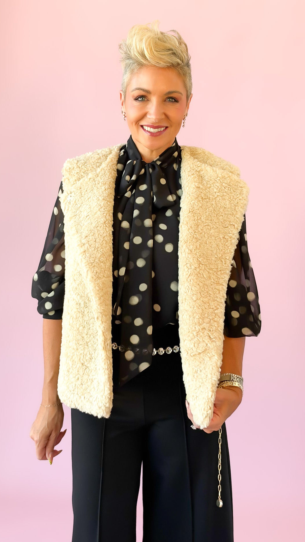 A woman modeling a cream sherpa vest over a black and white polka dot blouse with black wide-leg pants, standing against a solid pink background.