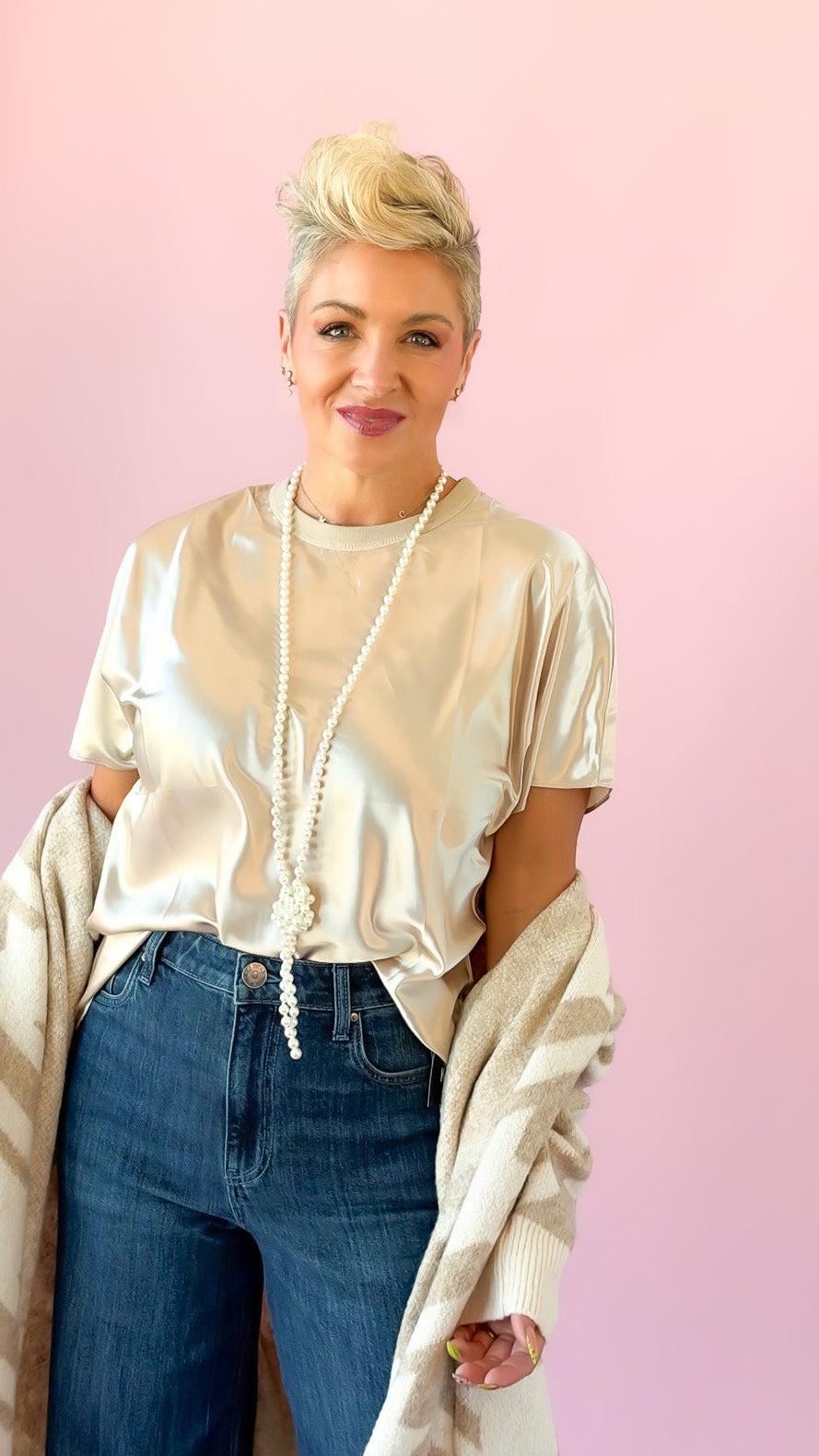 A woman modeling a cream satin short-sleeve top, blue wide-leg jeans, and a matching cream and gray patterned cardigan against a solid pink background.