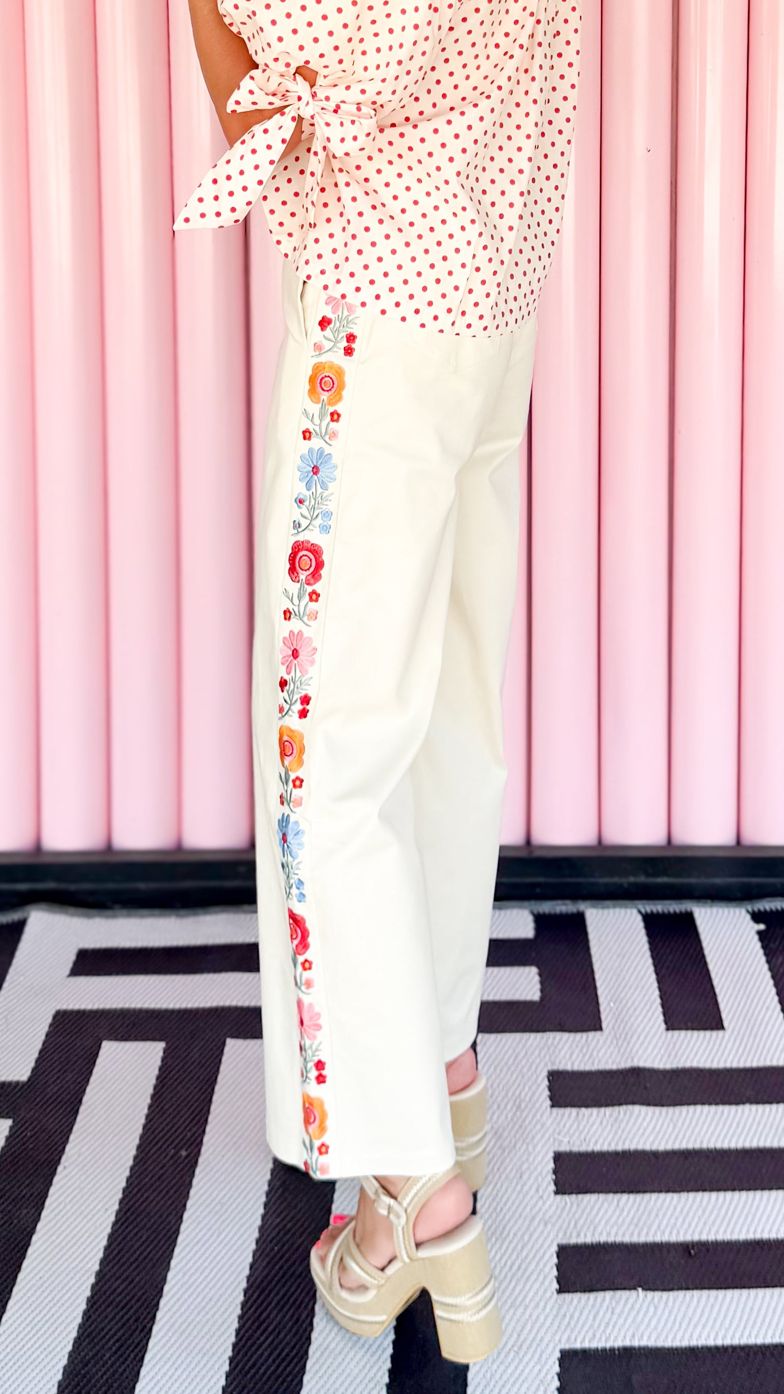 Woman in a polka dot top and floral pants standing against a pink striped wall.