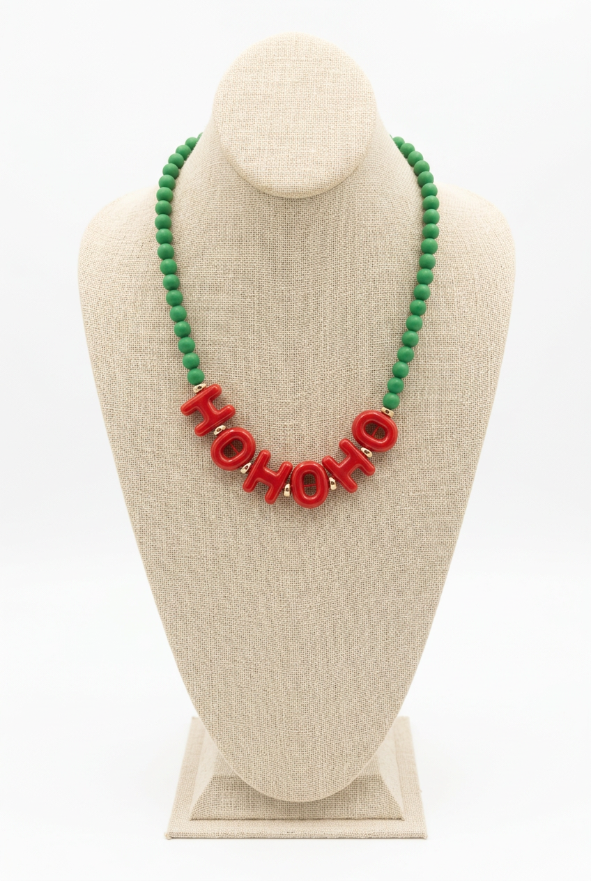 Green beaded necklace with red acrylic letters spelling HO HO HO at the center, displayed on a beige linen jewelry bust