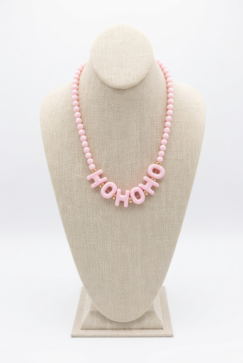 Pink beaded necklace with pink acrylic letters spelling HO HO HO at the center, displayed on a beige linen jewelry bust
