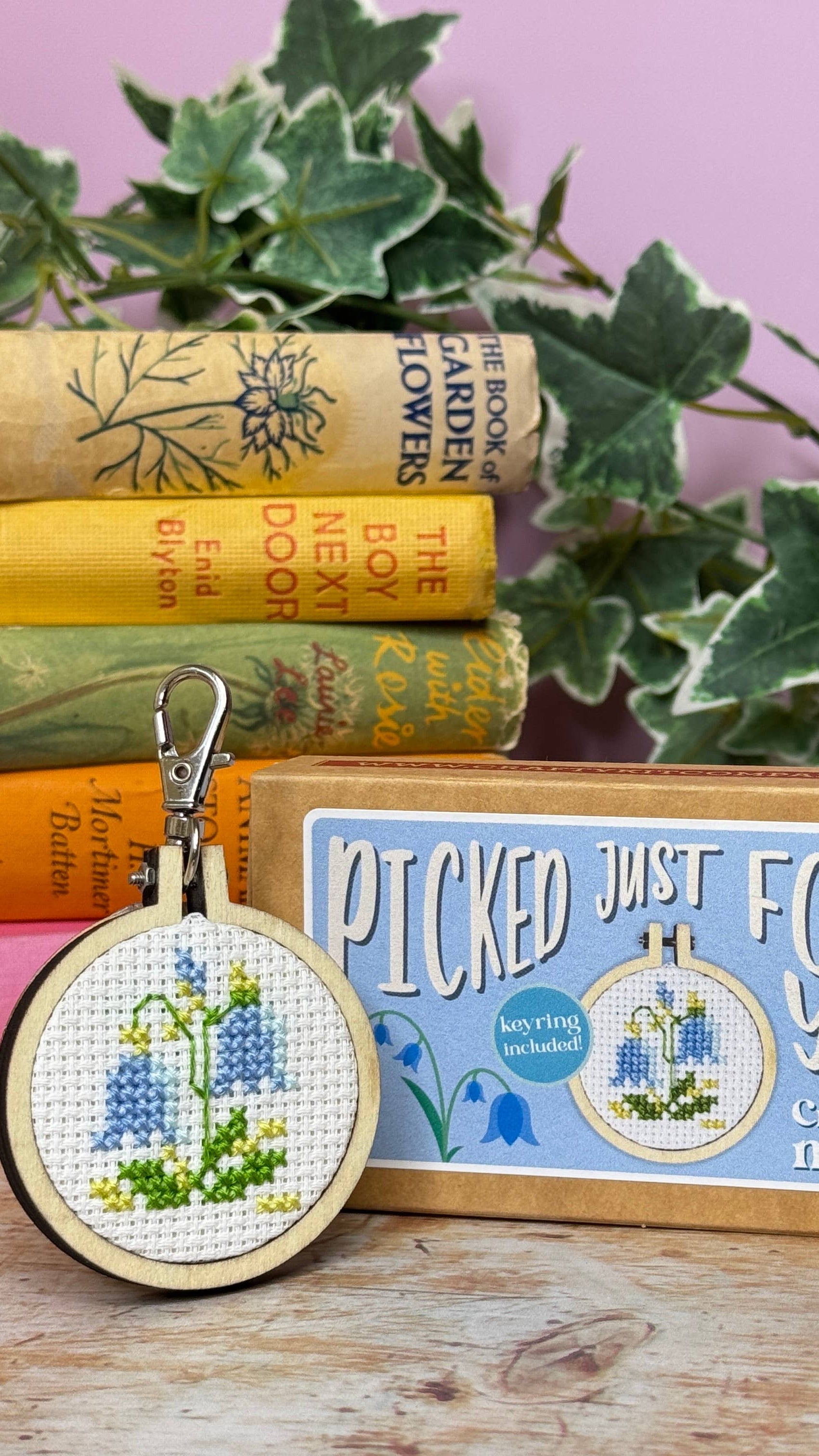 Cross-stitch mini kit with embroidery hoop and 'Picked Just For You!' text on a wooden surface with books and plants.