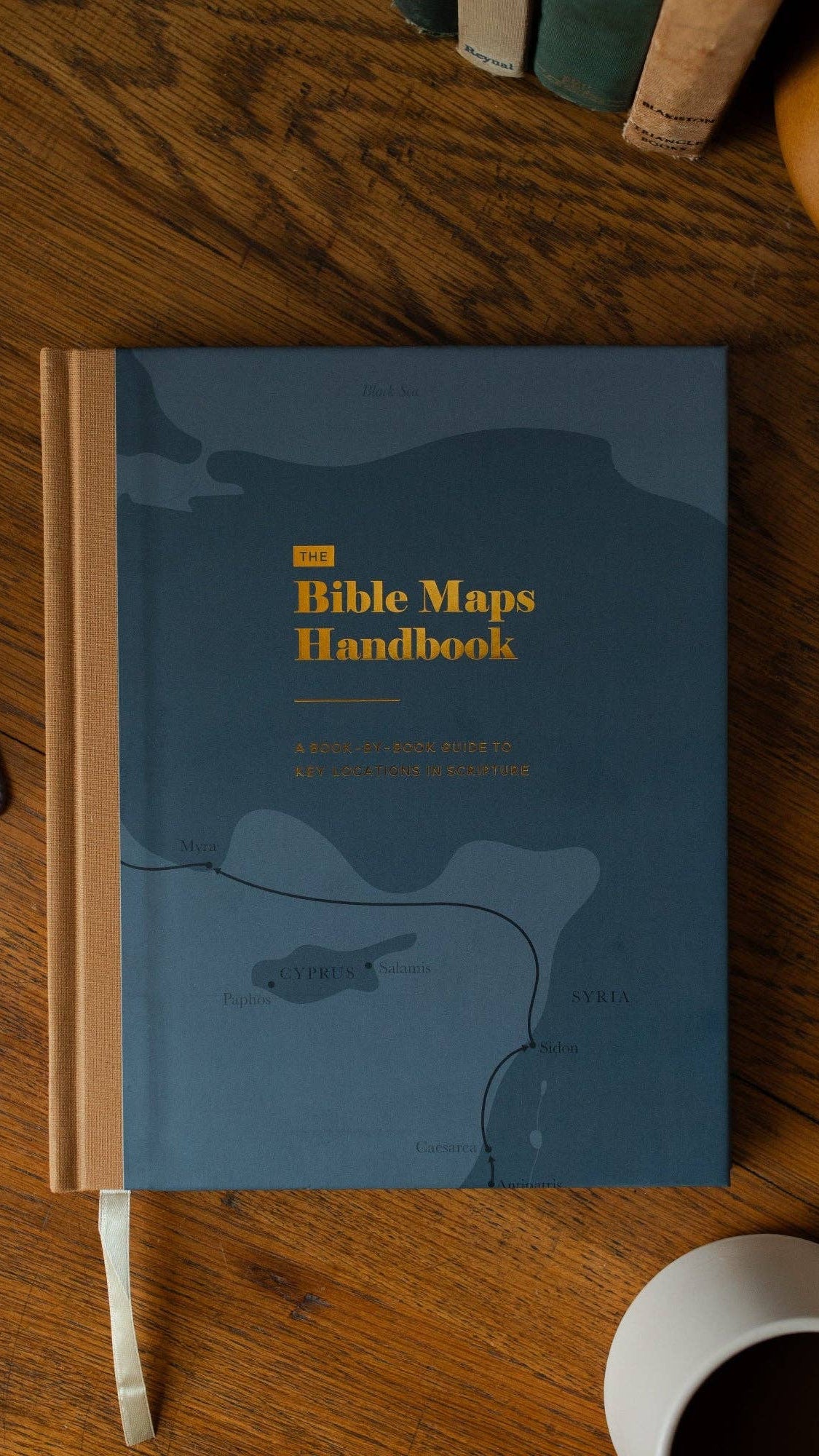 Blue book titled 'The Bible Maps Handbook' on a wooden surface with a cup of coffee.