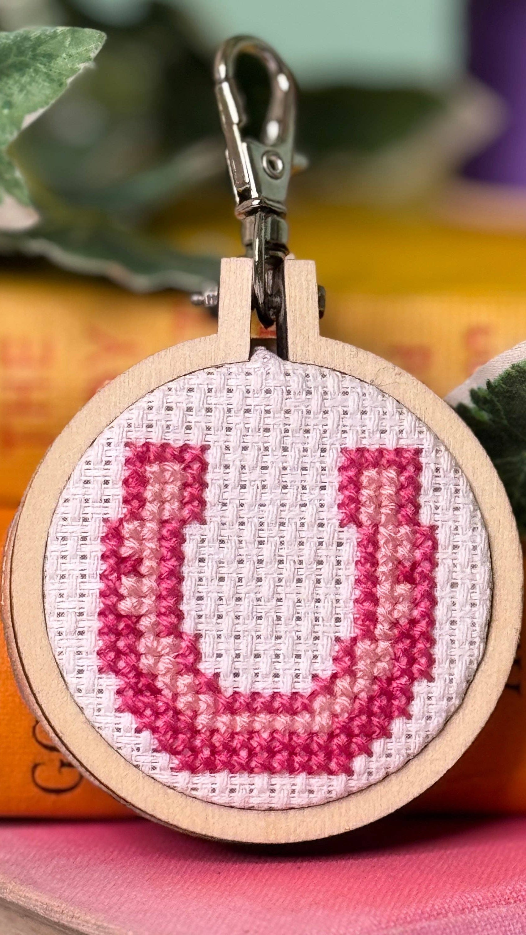 Cross-stitched letter 'U' keychain on a wooden hoop with books and leaves in the background
