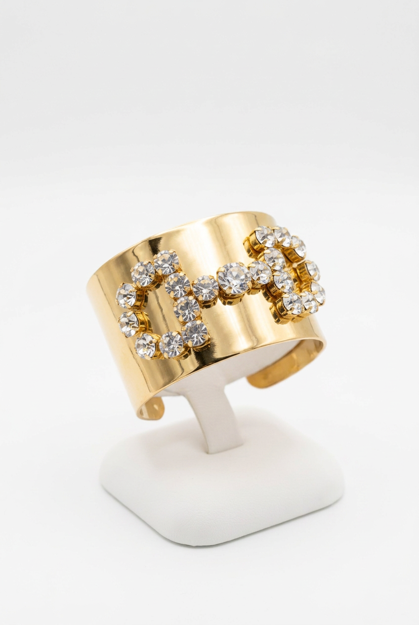 Wide polished gold-tone cuff with clear round stones arranged in a bow design on a white bracelet pedestal.
